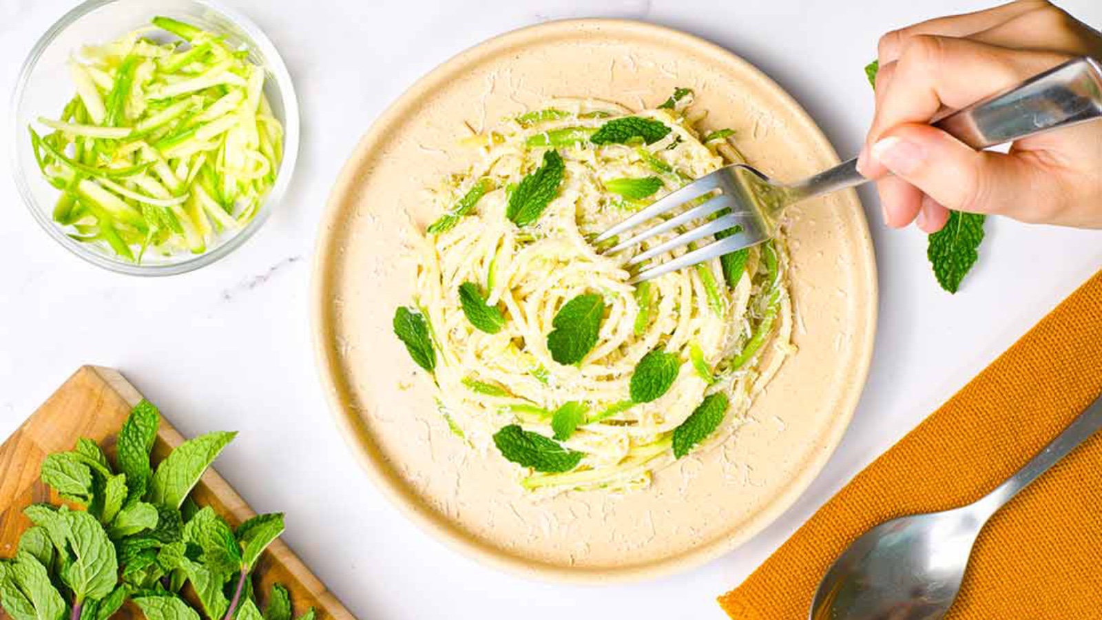 A hand with a fork reaches for some zucchini noodles on a plate.
