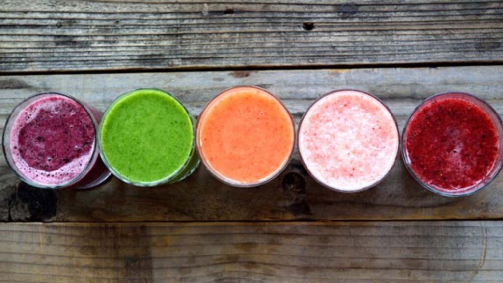 5 Smoothies in glasses on a wood surface, shown from overhead. Each smoothie is a different color.