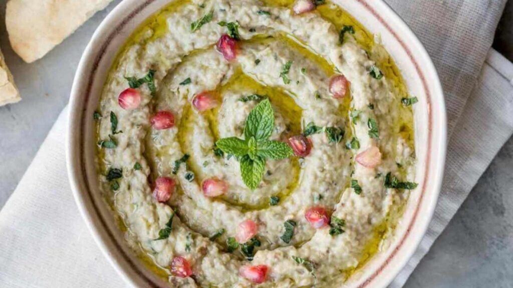 A bowl filled with baba ganoush and garnished with pomegranate arils and mint leaves.