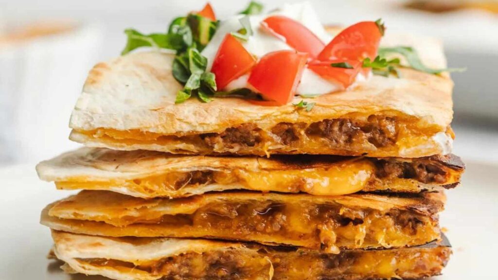 A stack of beef quesadilla topped with lettuce, sour cream and chopped tomatoes.