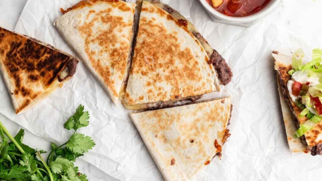 An overhead view of a black bean quesadilla on parchment paper. It has been cut into three pieces.