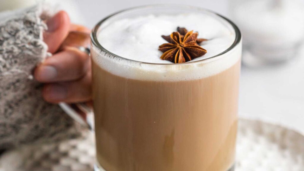 A hand holds a glass mug filled with dirty chai latte.