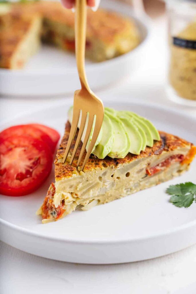 A slice of Spanish omelet on a plate with some tomato and avocado slices.