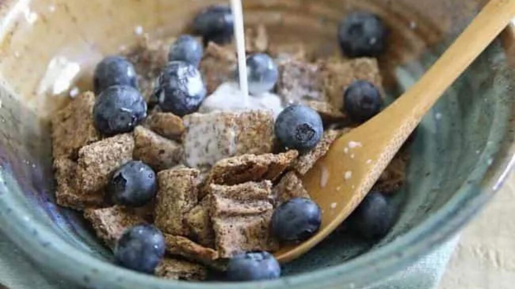 A bowl filled halfway with Homemade Cinnamon Crunch Cereal. A spoon rests in the bowl and some milk is being poured into it.