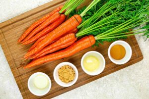 Fresh carrots with tops laying on a cutting board next to four small white bowls of recipe ingredients.