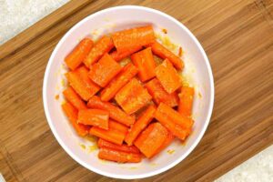 Carrots in a white mixing bowl mixed with seasoning and oil.