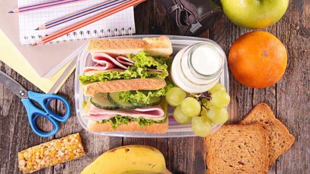An overhead view of a sandwich in a container with a small glass of milk and some grapes.