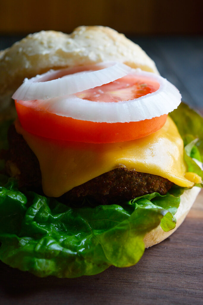 A turkey burger on a cutting board with the top bun slid to the back of the burger, showing the onions slices and tomatoes slices and cheese on top of the turkey patty.