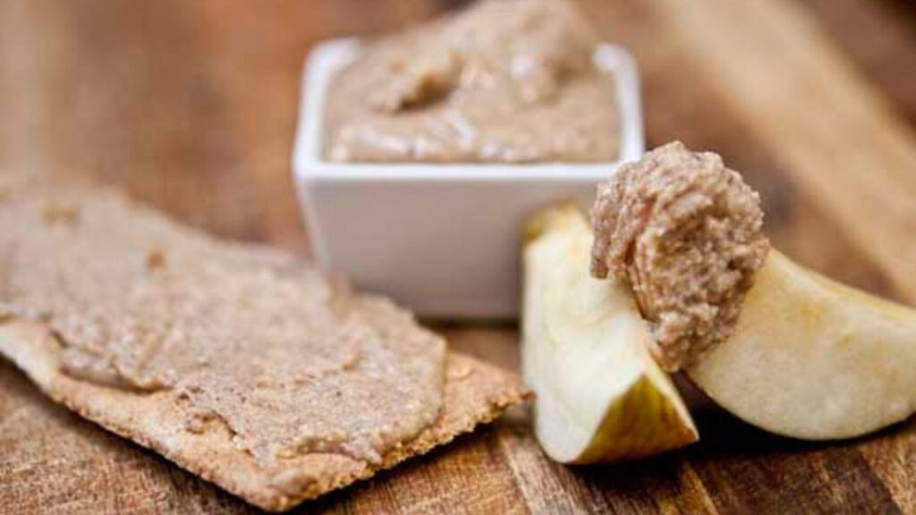 A cracker and apple wedge sit next to a small white bowl of almond butter.