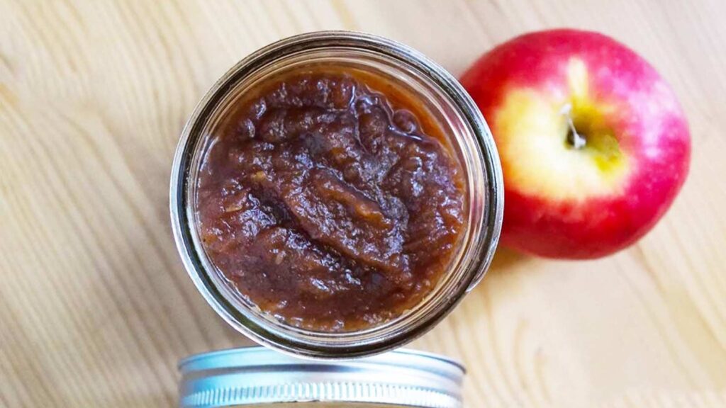 An overhead view of an open jar filled with amish apple butter. The jar lid and an apple lay to the sides of the jar.