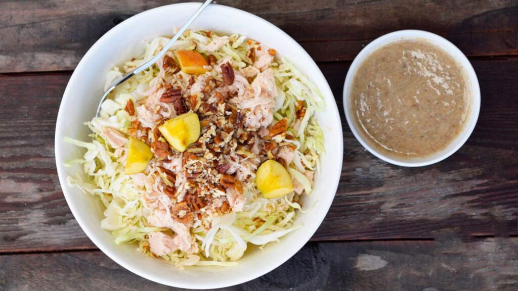 An overhead view of a Autumn Cabbage Salad in a white bowl. A small bowl of dressing sits to the right.