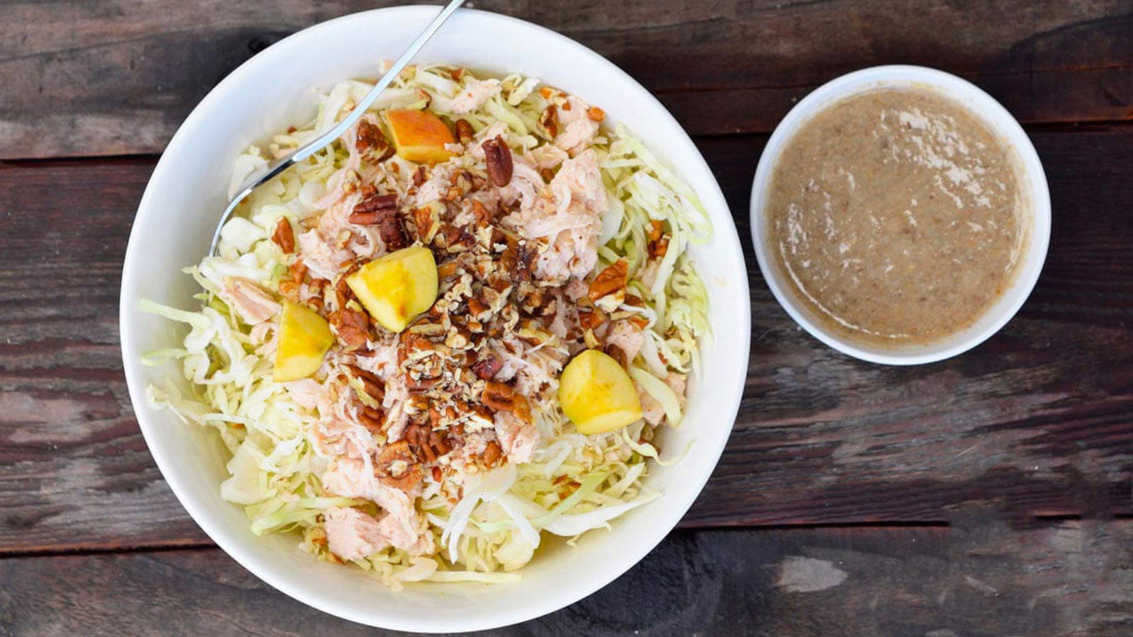 An overhead view of a Autumn Cabbage Salad in a white bowl. A small bowl of dressing sits to the right.