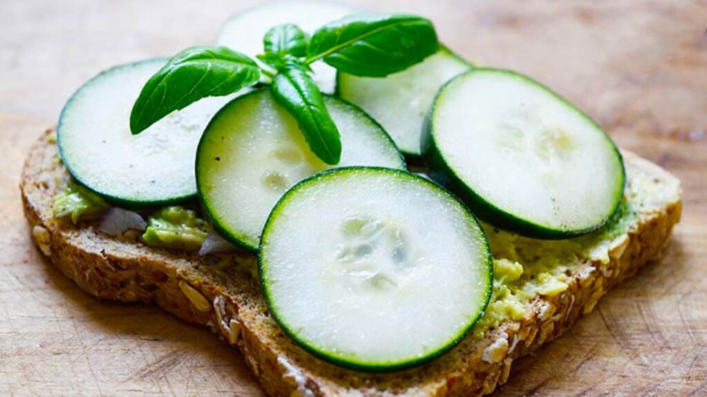 A slice of toast with mashed avocado, topped with slices of cucumber and garnished with fresh basil leaves.