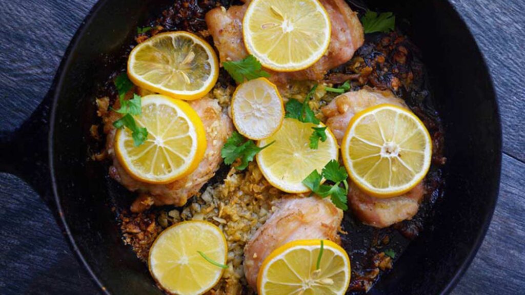 An overhead view of a black skillet filled with just baked garlic chicken thighs. Lemon slices lay on top of each chicken thigh.