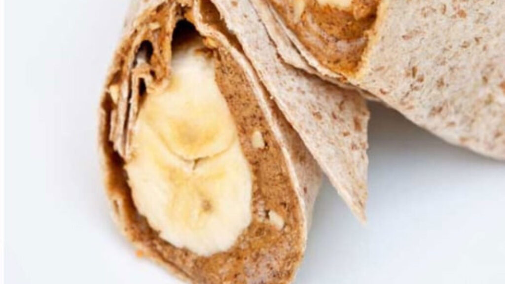 A cut banana wrap sits on a white background. You can see the layers of the whole grain wrap, nut butter and the bananas.