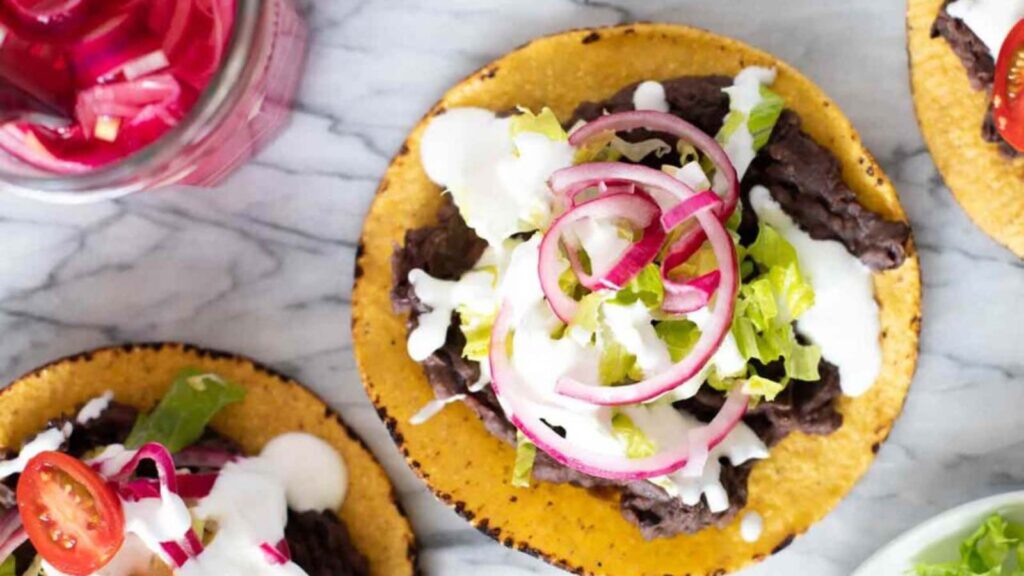 An overhead view of three black bean tostadas topped with lettuce, sour cream and onions.