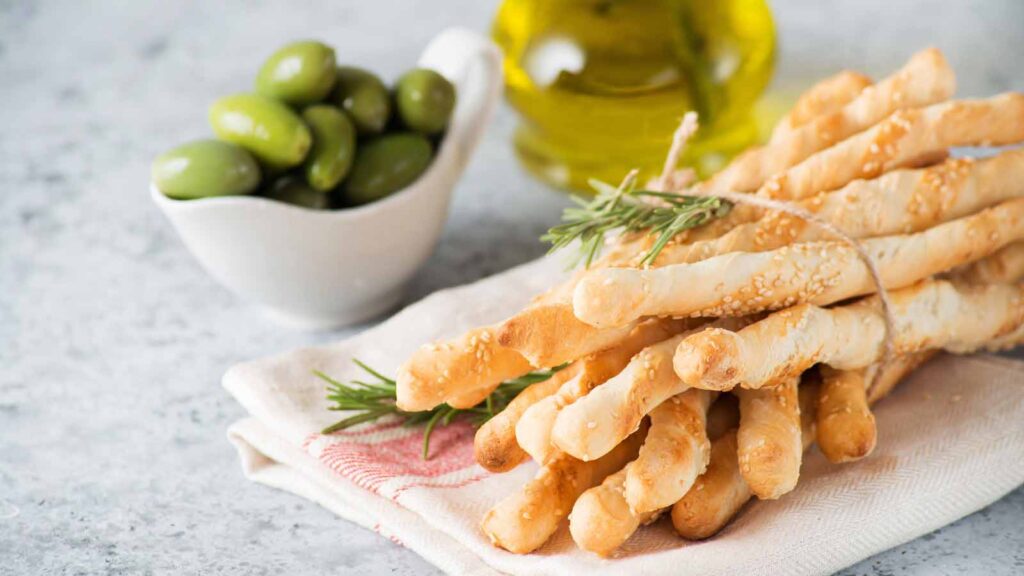 A bundle of breadsticks laying on a towel with a small bowl of olives and a jar of olive oil sitting behind it.