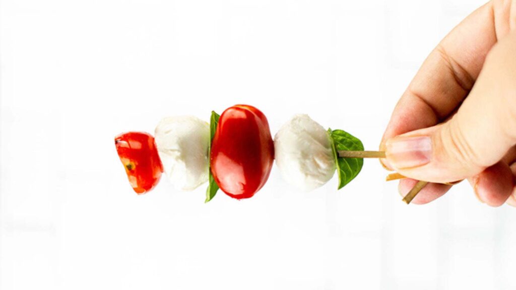 A hand holding a caprese skewer against a white background.