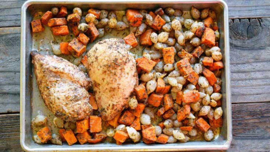 A sheet pan covered in sweet potato chunks, pearl onions and two chicken breasts.