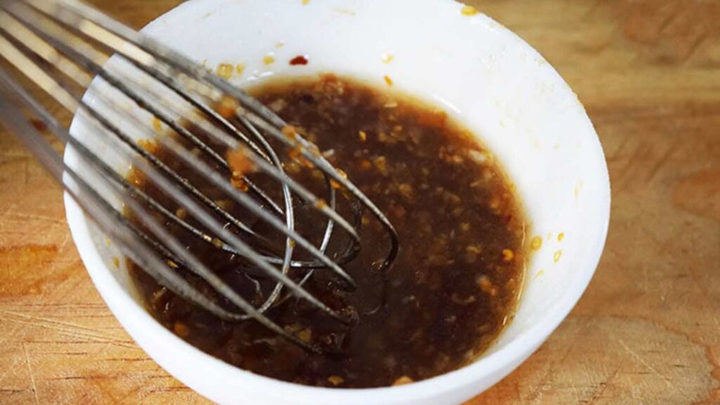 A whisk resting in a small white bowl which holds some Chinese Garlic Sauce.