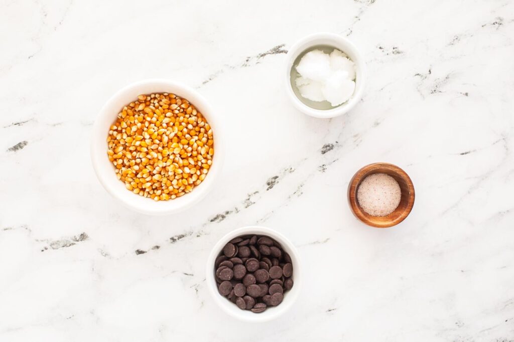 Chocolate Popcorn Recipe ingredients in individual bowls on a white marble surface.