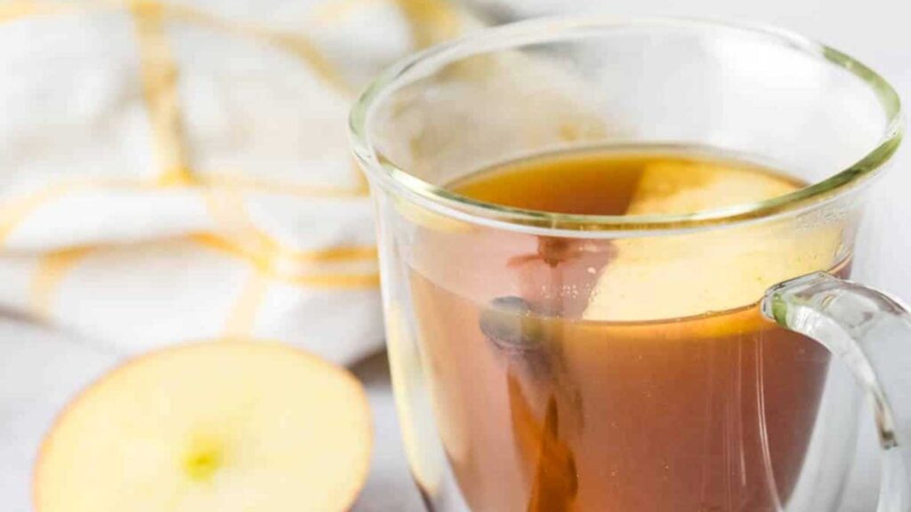 A closeup of a glass mug filled with apple cider tea with a slice of apple floating in it.