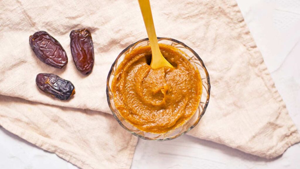 The finished date caramel in a clear glass serving bowl with a small wooden spoon for serving, resting in the bowl.