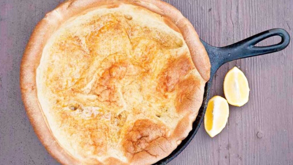 An overhead view looking down into a cast iron pan with a dutch baby pancake in it. Two lemon wedges sit to the right of the skillet.