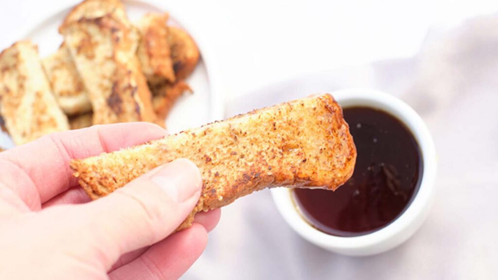 A female hand dips a French Toast Stick into a small cup of maple syrup.