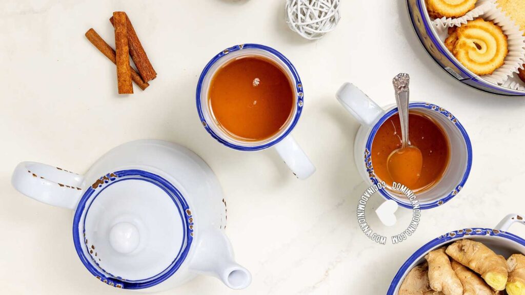 An overhead view of two tea mugs and a tea pot on a table next to a container of fresh ginger and another container of holiday cookies.