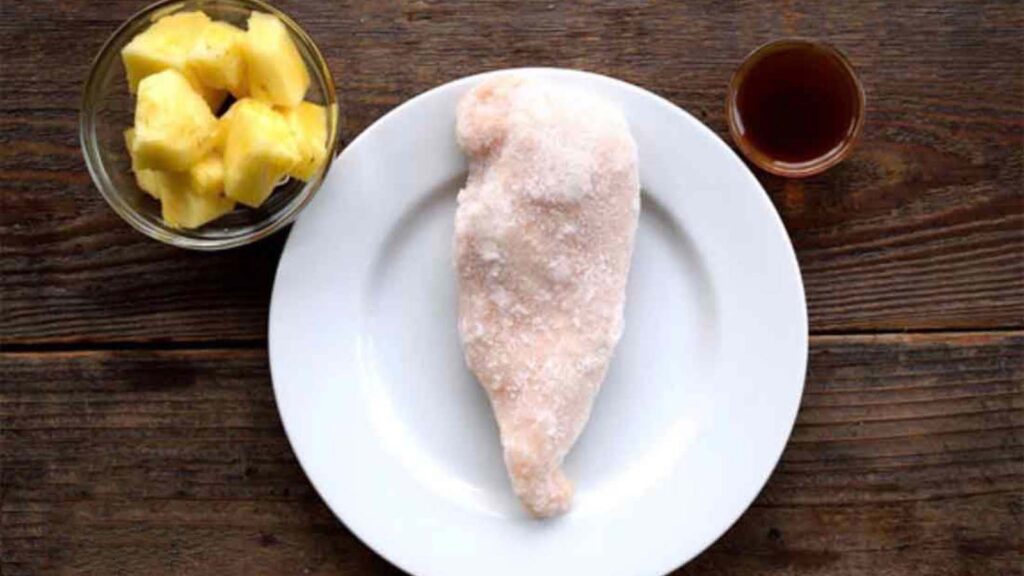 A frozen chicken breast on a white plate sits on a wood surface. Two small bowls sit on either side of it, one filled with pineapple chunks and the other with a brown sauce.