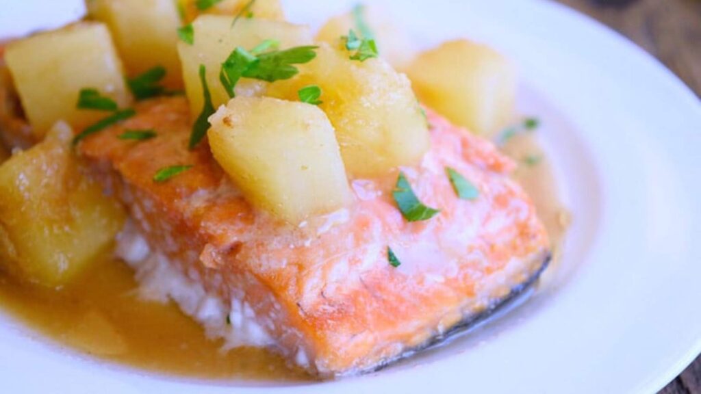 A Hawaiian salmon filet on a white plate, topped with pineapple chunks and chopped herbs.