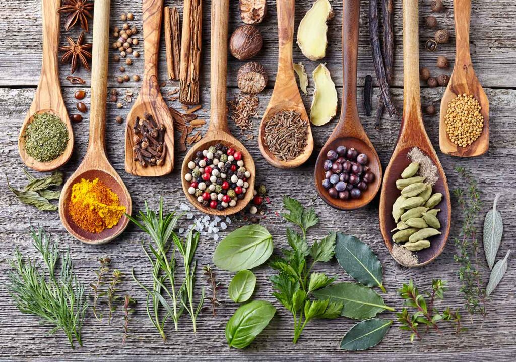 Wooden spoons lined up on a wood surface, holding dried herbs and spices. Fresh herb leaves lay in front of the spoons.