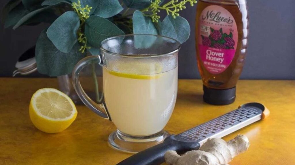 A glass mug filled with honey lemon ginger tea. A jar of honey, a half lemon and a grater with some fresh ginger lay next to the mug.