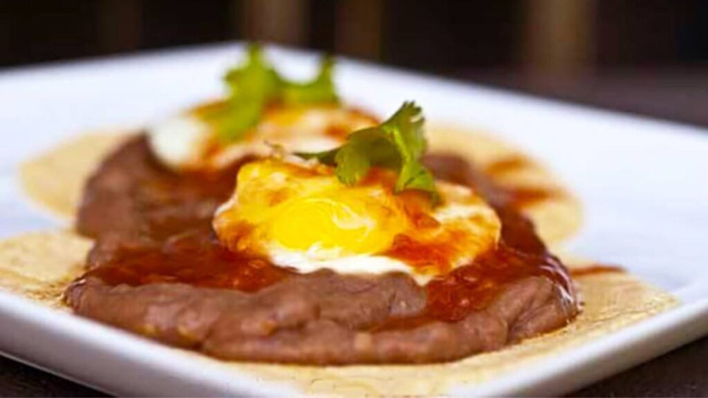 A square, white platter with huevos ranchers on it. A corn tortilla with refried beans and an over medium egg on top.