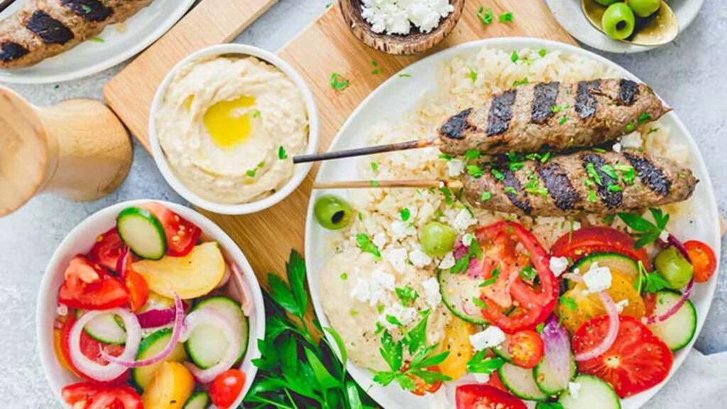 Beef Kafta laying on a bed or rice with fresh sliced tomatoes and cucumbers next to them.