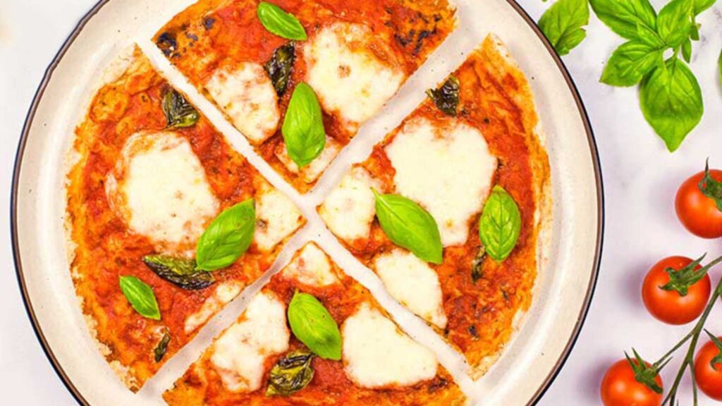 A sliced Margherita Pizza on a plate. Fresh basil and cherry tomatoes on the stem lay to the right of the plate.