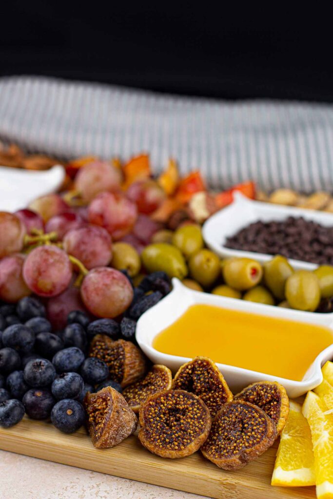 A center front view of a Dessert Mezze Platter shows two bowls of dip, dried figs, fresh blueberries, grapes, olives, and more.