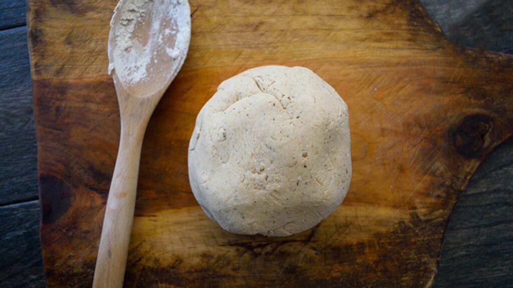 A ball of pizza dough on a cutting board. A used wooden spoon lays next to it.