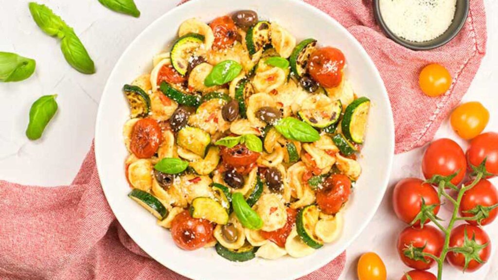 A white bowl filled with Orecchiette Pasta Salad sitting on a red dishtowel, surrounded by fresh basil leaves and cherry tomatoes.