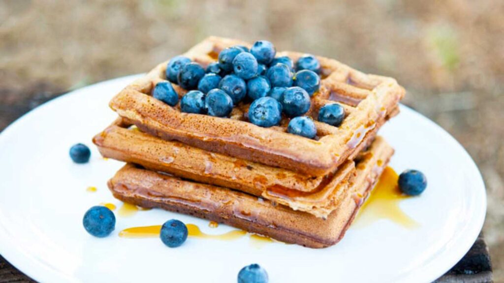 A white plate holds a stack of three homestyle waffles that are topped with fresh blueberries.