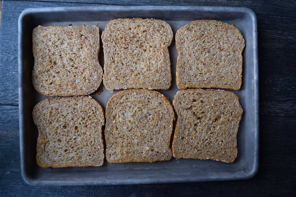 Six slices of whole grain bread on a baking sheet.