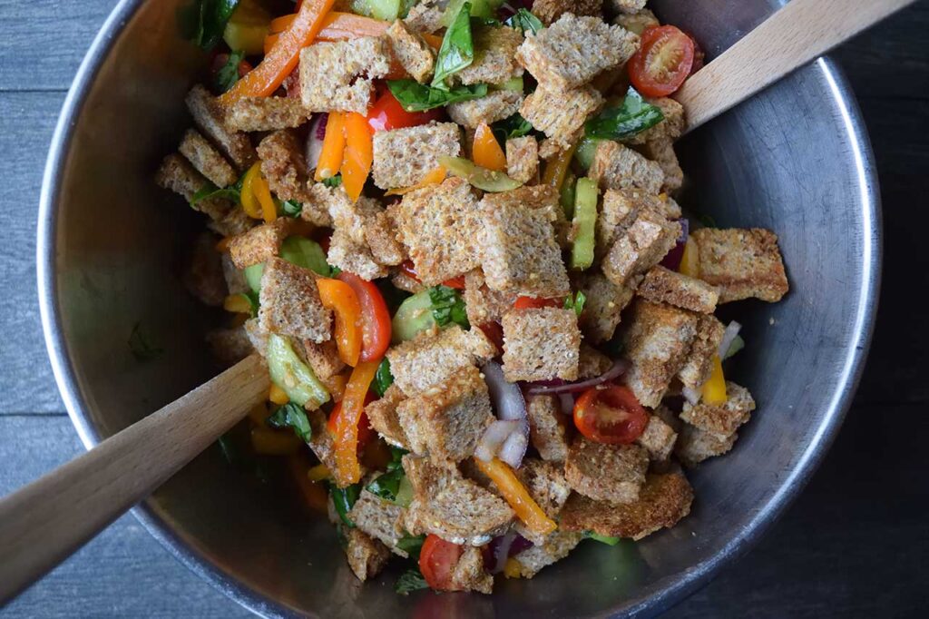 Panzanella mixed together in a mixing bowl. Wood tongs rest in the bowl.