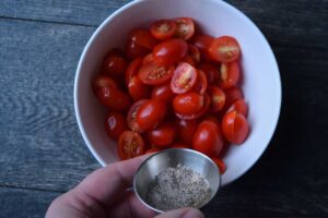Adding slat and pepper to grape tomatoes in a white bowl.