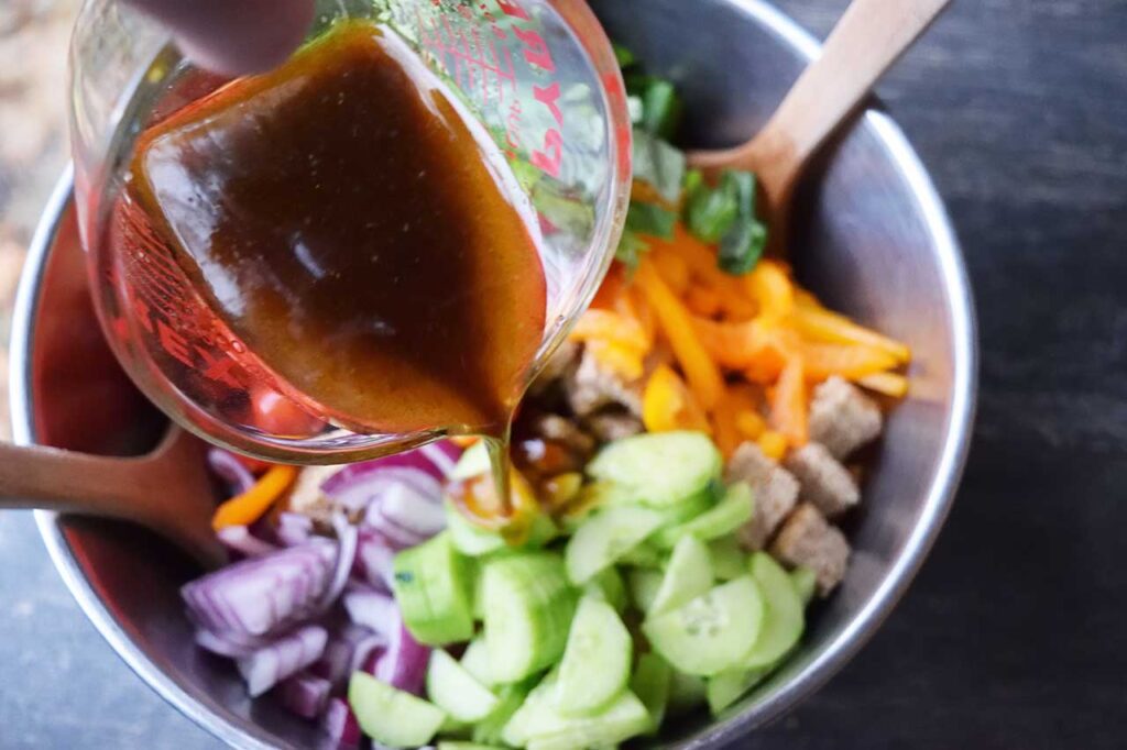 Pouring vinaigrette over the Panzanella ingredients in a large mixing bowl.
