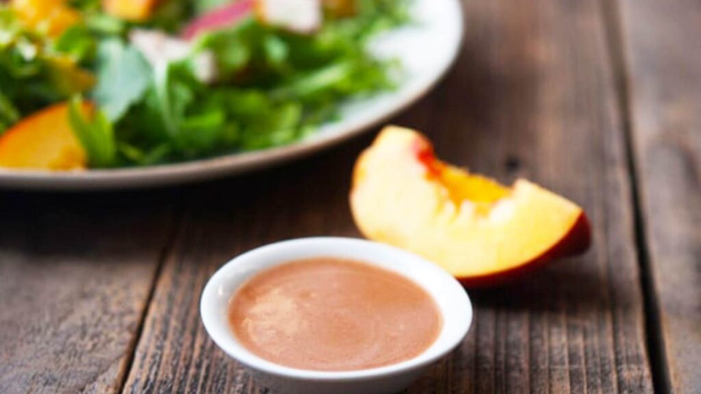 A small, white dish filled with Peach Vinaigrette next to a slice of fresh peach and a plate with peach salad.
