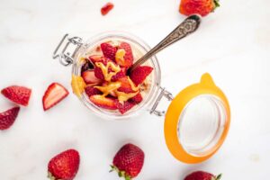 An open jar with Peanut Butter and Jelly Overnight Oats and a spoon on a white surface with fresh strawberries laying around it.