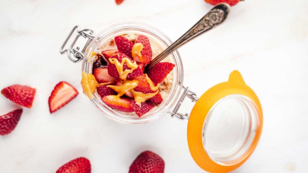 An open jar with Peanut Butter and Jelly Overnight Oats and a spoon on a white surface with fresh strawberries laying around it.