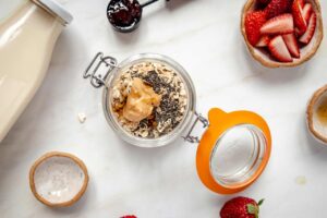 Peanut butter added to oats and seeds in an open canning jar on a white surface.