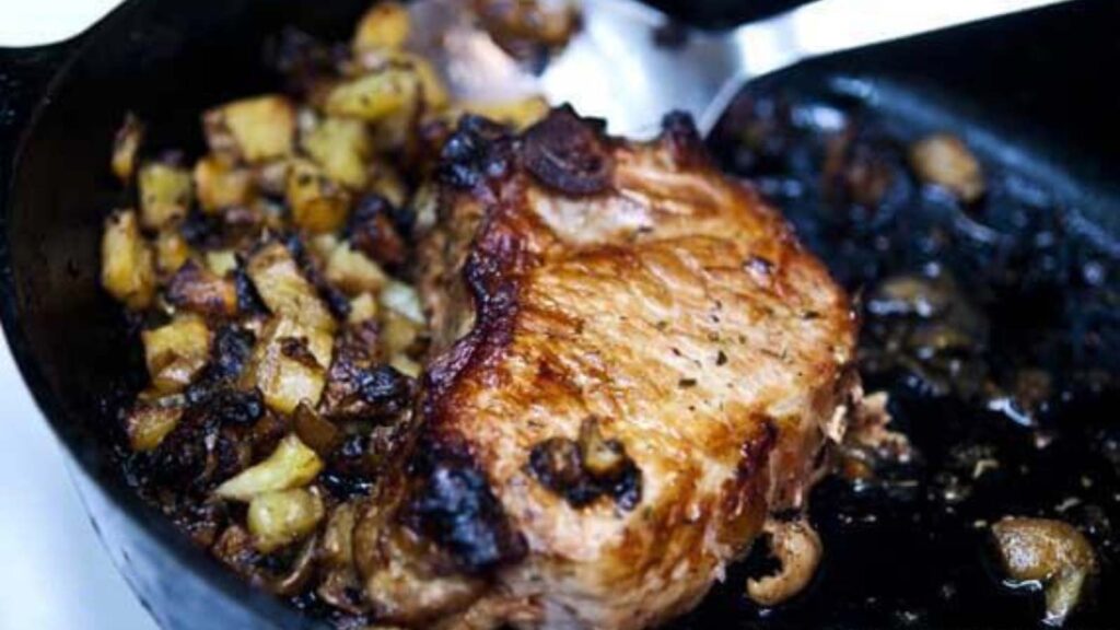 A black skillet with a pork chop and fried potatoes in it.